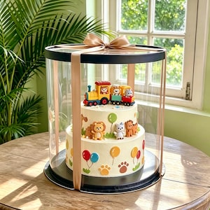 May include: A two-tier white frosted cake inside a clear cylindrical display box with a gold ribbon bow on top. The cake is decorated with colorful fondant animal figures, balloons, and paw prints, topped with a toy train carrying animal passengers.
