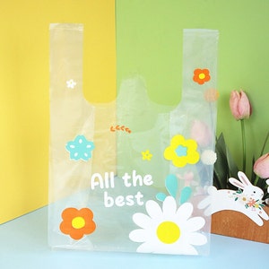 Puede incluir: Una bolsa de pl&aacute;stico transparente con un dise&ntilde;o floral. La bolsa tiene una margarita blanca con un centro amarillo, una estrella azul y flores naranjas y amarillas. El texto "All the best" est&aacute; impreso en la bolsa.