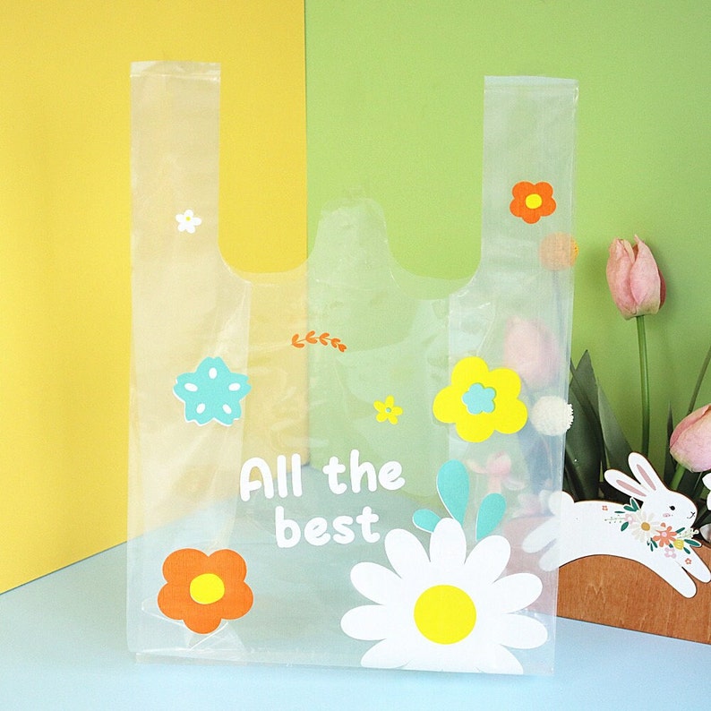 Puede incluir: Una bolsa de pl&aacute;stico transparente con un dise&ntilde;o floral. La bolsa tiene una margarita blanca con un centro amarillo, una estrella azul y flores naranjas y amarillas. El texto "All the best" est&aacute; impreso en la bolsa.