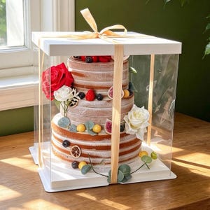 May include: A three-tiered naked cake inside a square clear plastic box with a white base and lid, tied with a light beige ribbon. The cake is decorated with fresh red and white roses, sliced figs, blueberries, strawberries, and dried citrus.