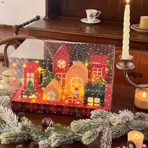 May include: A Christmas village advent calendar with a clear plastic cover. The houses are made of paper and have red, green, and brown accents. The houses are lit with small white lights. The calendar is sitting on a wooden table with a plaid pattern.