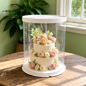 May include: A two-tier cream-colored cake decorated with pink and peach-toned frosting flowers and green leaves, topped with a small tiger figurine, enclosed within a clear cylindrical display case with a white base and lid.