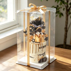 May include: A white cylindrical cake with gold drip icing, topped with a gold crown, black and gold star cutouts, and a clear balloon with the text Happy Birthday. The cake is encased in a tall, clear plastic box tied with a gold ribbon.