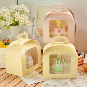 May include: Three clear-windowed cake boxes with handles. The boxes are yellow and pink. The boxes are empty, but there are images of cakes inside the windows.