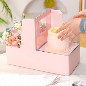 May include: A pink box with a clear plastic window, containing a small pink and yellow cake. The box also has a separate compartment for flowers.
