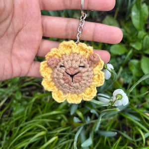 May include: A handmade crochet keychain featuring a lion's face. The lion has a yellow mane, a tan face, and closed eyes. The keychain is attached to a silver metal ring and chain. The background is blurred with green grass and white flowers.