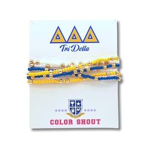 May include: A set of three beaded bracelets in the colors of Tri Delta sorority: white, gold, and blue. The bracelets are displayed on a white card with the sorority's name and logo. The card also says "Color Shout".