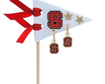 May include: A white triangular pennant display card with two red ribbons, featuring a red and black NC logo and two gold star-shaped studs. Two gold hoop earrings with small NC logo charms hang from the card, each accented with clear crystals.