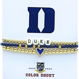 May include: Three beaded bracelets. One bracelet spells "DUKE" with white letter beads. The other two bracelets are gold and navy blue. A gold heart with a blue center is on one bracelet. The Duke logo and "COLOR SHOUT" are also visible.