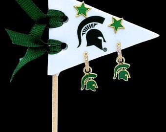 Könnte beinhalten: Goldfarbene Ohrringe mit grünen Spartanerhelm-Anhängern und kleinen, klaren Steinen. Die Ohrringe werden auf einem weißen Wimpel mit einem grünen Band und zwei grünen sternförmigen Ohrsteckern präsentiert. Der Wimpel hat ein Spartanerhelm-Logo.