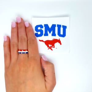 May include: A hand wearing a red, white, and blue ring with gold accents. The ring has white dots on the red and blue bands. The hand is holding a sticker with the text "SMU" in blue letters and a red horse running to the right.