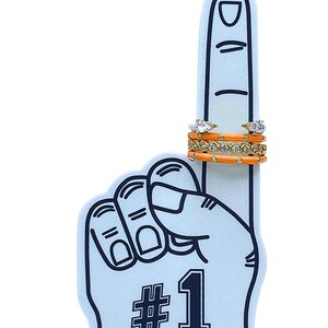 May include: A light blue foam finger with the number 1 in black. Two rings with clear stones and an orange enamel band are displayed on the finger. A gold band ring with clear stones is also shown.