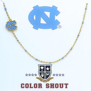 May include: A gold-colored necklace with a light blue enamel charm featuring the North Carolina Tar Heels logo. The necklace has small blue beads and is displayed on a white background with the "Color Shout" logo.