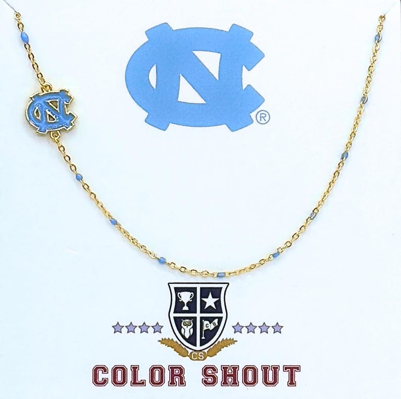 May include: A gold-colored necklace with a light blue enamel charm featuring the North Carolina Tar Heels logo. The necklace has small blue beads and is displayed on a white background with the "Color Shout" logo.