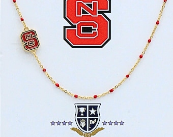 May include: A gold-tone chain necklace featuring small red enamel beads spaced along the links and a rectangular charm displaying the red and white NC logo. The design includes a larger NC logo graphic above and a shield emblem with the text COLOR SHOUT.