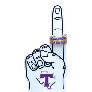 May include: Three rings are displayed on a foam finger. The rings include a gold band with clear stones, a purple band with gold dots, and a gold band with pear-shaped stones. The foam finger is white with a purple and white logo.