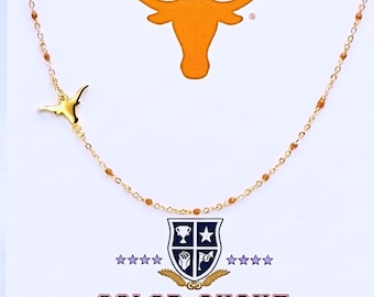 May include: A gold-toned chain necklace featuring small brown beads and a gold longhorn steer pendant. The necklace is displayed on a white card with an orange longhorn logo, a shield emblem, and the text COLOR SHOUT printed in red letters.