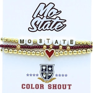 May include: A set of three beaded bracelets on a display card labeled Mo State and Color Shout. The bracelets feature gold-tone metallic beads, maroon seed beads, and white letter beads spelling MO STATE, with a central red heart charm.