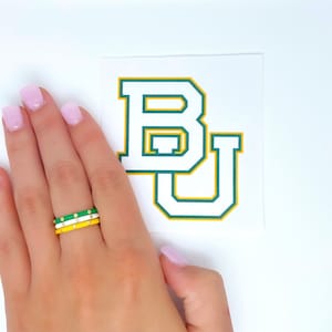 May include: A hand wearing three stacked rings. The rings are white, yellow, and green with gold accents. The rings are on a finger with pink nail polish. The background is a white surface with a green and yellow "BU" logo sticker.