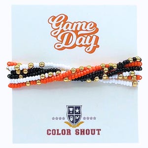 May include: A set of beaded bracelets in white, black, and orange, accented with gold-colored beads. The bracelets are displayed on a white card with the words "Game Day" in orange script and the text "Color Shout".