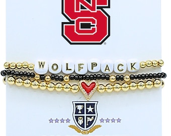 May include: A set of three beaded bracelets on a white display card featuring the NC State logo. One bracelet has white letter beads spelling WOLFPACK, one has small black beads, and one has gold beads with a central red heart charm with crystals.