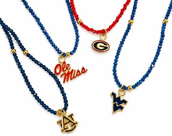 May include: Four beaded necklaces featuring small faceted blue or red beads accented with gold-tone spacers. Each necklace displays a unique metal charm, including the red script text Ole Miss, a gold and black G logo, a gold AU logo, and a blue WV logo.