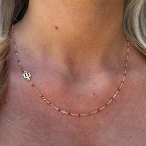 May include: A gold-colored necklace with a red beaded chain and a gold-colored pendant. The pendant features a stylized trident design. The necklace is worn around the neck.