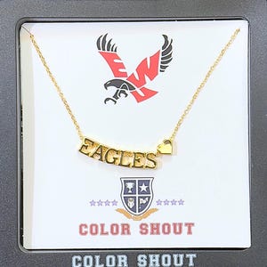 May include: Gold-toned necklace with the word "EAGLES" and a heart charm. The necklace is displayed on a white card with a red and black eagle logo and the text "COLOR SHOUT". The packaging is black.