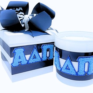 May include: A white gift box with a blue and white striped design and a blue bow. The box is labeled with the letters "A A O". Next to the box is a white candle with a blue and white striped design and the letters "A A O" on the front.