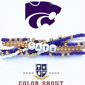 May include: A set of purple, white, and gold beaded bracelets with the word "CATS" spelled out in white beads. The bracelets are layered on top of each other. The image also includes a purple mascot of a wildcat and a logo for "COLOR SHOUT" with a shield and stars.