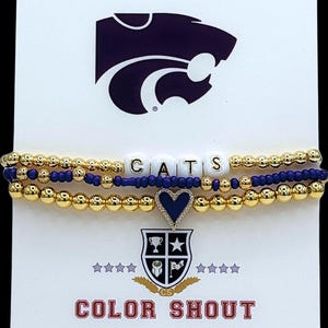 May include: Three-strand bracelet set with gold beads, navy blue beads, and white letter beads spelling "CATS." A navy blue heart charm and a shield with a trophy and star are included. The text "COLOR SHOUT" is at the bottom.