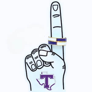 May include: A white foam finger with a black outline, pointing upwards, with a ring on the index finger. The ring has a gold band with white and purple enamel. A purple "T" logo is on the palm.
