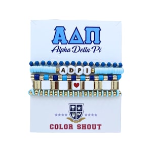 May include: A set of beaded bracelets in shades of blue, white, and gold, displayed on a white card. The bracelets feature letter beads spelling "ADPI" and a red heart bead. The card has the text "Alpha Delta Pi" and "COLOR SHOUT".
