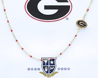 May include: A gold-tone chain necklace featuring small red enamel accents and a single oval charm with a black and gold Georgia G logo. The necklace is displayed on a white card printed with a large Georgia G logo and the text COLOR SHOUT.