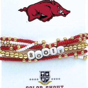 May include: Stackable beaded bracelets in red, white, and gold. The bracelets spell out "SOOIE" with white letter beads. A red Razorback logo is at the top, with "COLOR SHOUT" printed at the bottom.