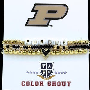 May include: Three beaded bracelets. The top bracelet spells out "PURDUE" with white letter beads. The other two bracelets are gold and black. A gold heart charm with a black center hangs from the bottom bracelet. The Purdue logo and "COLOR SHOUT" text are visible.