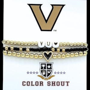 May include: Three beaded bracelets. One gold, one black, and one with white letter beads spelling "VU" and a heart. The bottom bracelet has a black heart charm. The top of the card has a gold "V" logo. The bottom of the card says "COLOR SHOUT".