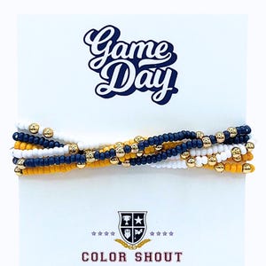 May include: A stack of beaded bracelets in white, navy blue, and gold, with gold accent beads. The bracelets are displayed on a white card with the text "Game Day" in navy blue script and the words "Color Shout" below.