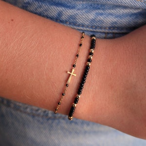 May include: Two bracelets on a wrist. One bracelet is a gold chain with a small gold cross charm. The other bracelet is black beads with gold accents.