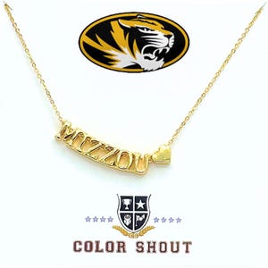 May include: A gold-colored necklace with the word "MIZZOU" and a heart charm. The necklace is displayed on a white background with the "Mizzou" logo and the text "COLOR SHOUT".