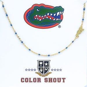 May include: A gold-tone chain necklace featuring small blue enamel accents along the links and a gold-tone letter F charm. The necklace is displayed on a white card featuring a green alligator logo and the text COLOR SHOUT with a crest emblem.