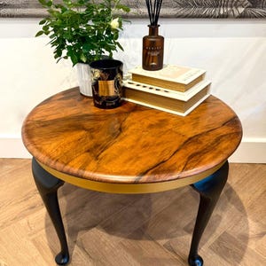 May include: A circular coffee table with a dark wood grain top, black legs, and a gold trim. The table supports a plant, books, a candle, and a diffuser. The table's diameter is approximately 76 cm.