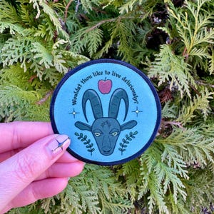 May include: A blue and black round patch with a goat head illustration and the text "Wouldst thou like to live deliciously?" with a red apple above the goat's head.