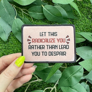 May include: A rectangular patch with the text "LET THIS RADICALIZE YOU RATHER THAN LEAD YOU TO DESPAIR" in black letters. The patch has a light tan background and a black border. The patch is held by a hand with green nail polish.