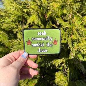 May include: Green patch with black border and white text that reads "seek community amidst the chaos".  Two red flames are on either side of the text.