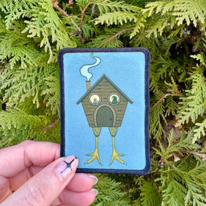 May include: A blue fabric patch with a brown house with two windows and a door. The house has legs and is standing on a blue background. There is smoke coming from the chimney.