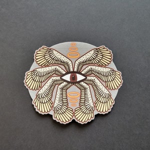 May include: A circular sticker with a stylized eye surrounded by eight wings. The wings are a light yellow with brown outlines. The eye is brown with a white pupil. There are four orange rings above the eye.