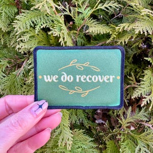 May include: Green fabric patch with a black border and white text that reads "we do recover" surrounded by a gold floral design.