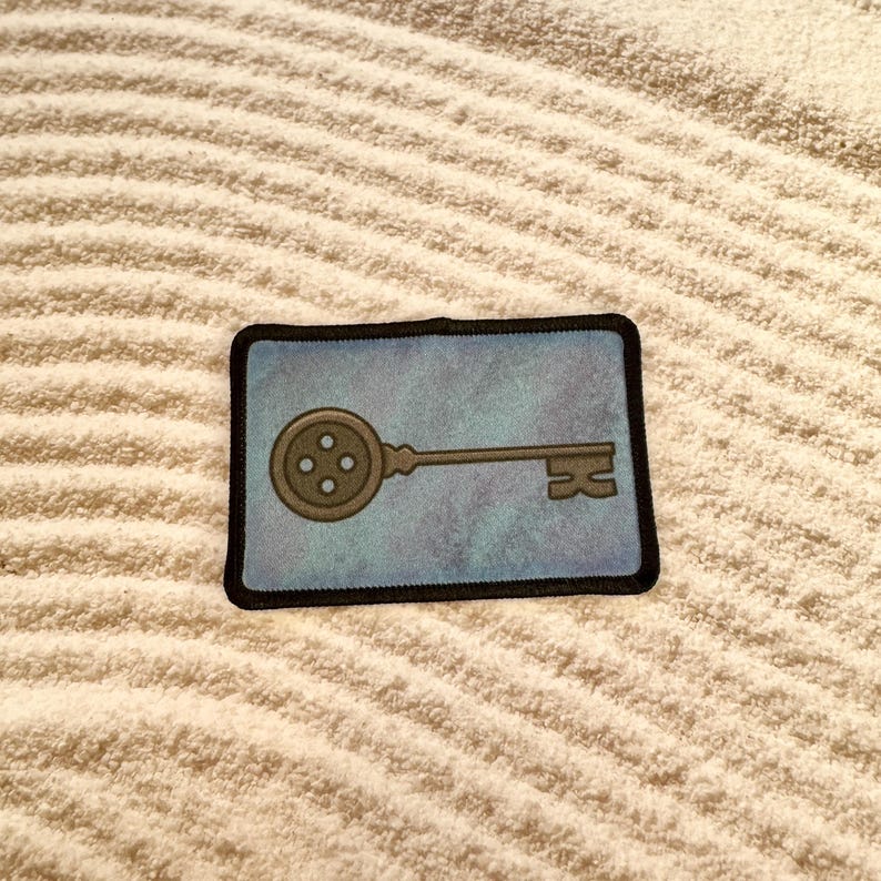 May include: A blue and gray embroidered patch with a keyhole shaped key with a button for a head. The key is on a blue background with a white border.
