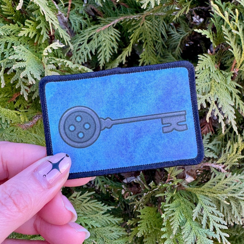 May include: A blue fabric patch with a black outline, featuring a key with a button-shaped head. The key is a dark gray color.